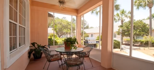 Screened-in porch with a round glass table, four metal chairs, potted plants, and views of palm trees and neighboring houses.