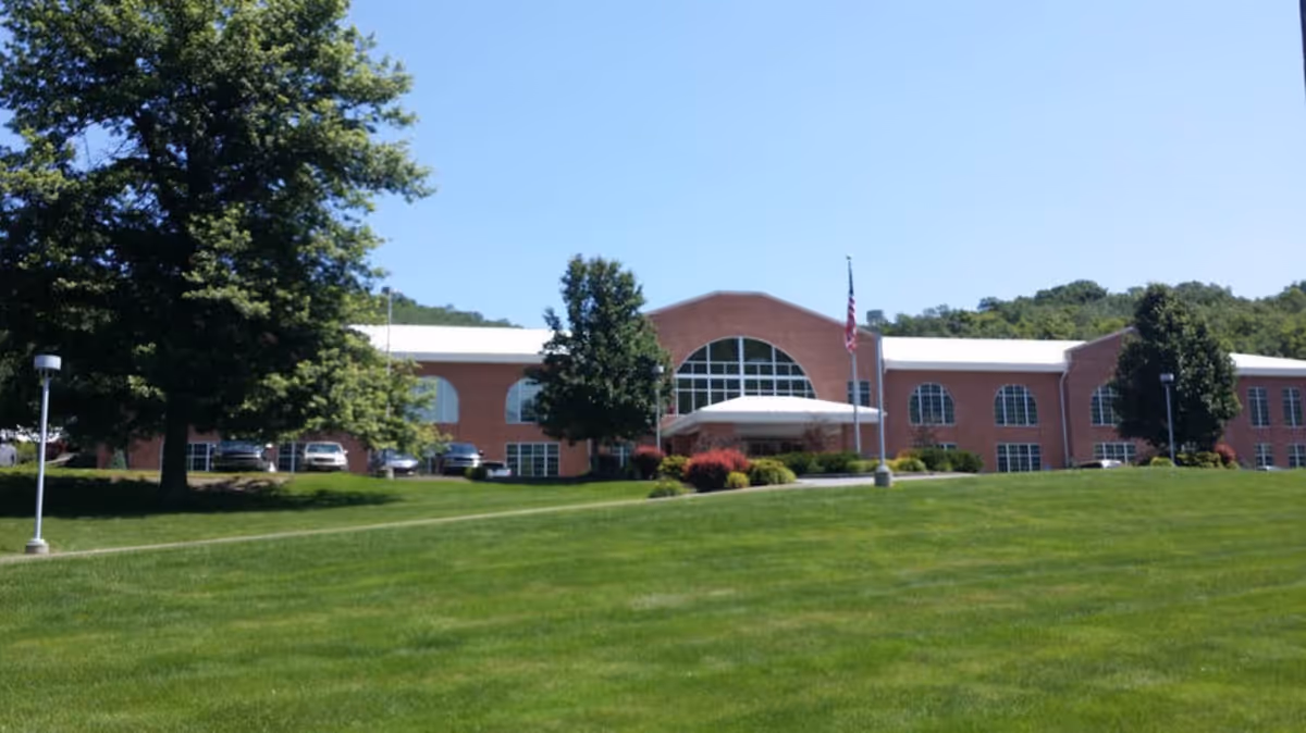 A large brick building with multiple arched windows, surrounded by green grass and trees under a clear blue sky. There is an American flag on a flagpole near the entrance, and several cars are parked along the side of the building.