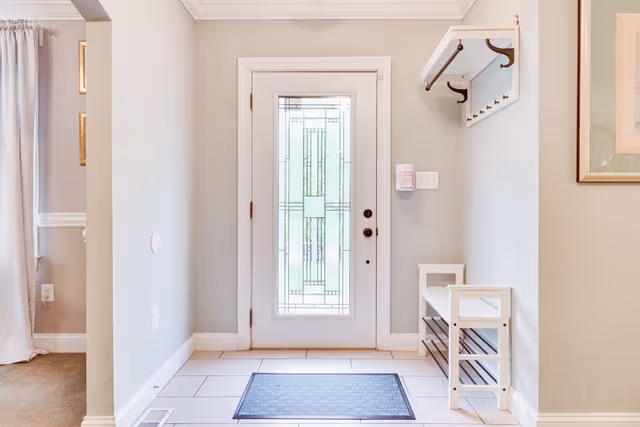 A bright entryway with a white door featuring decorative glass panels. The floor is tiled with a small dark mat in front of the door. To the right, there is a white wall-mounted coat rack with hooks and a small white bench with shelves underneath. Light gray walls and a framed picture are visible on the right side.