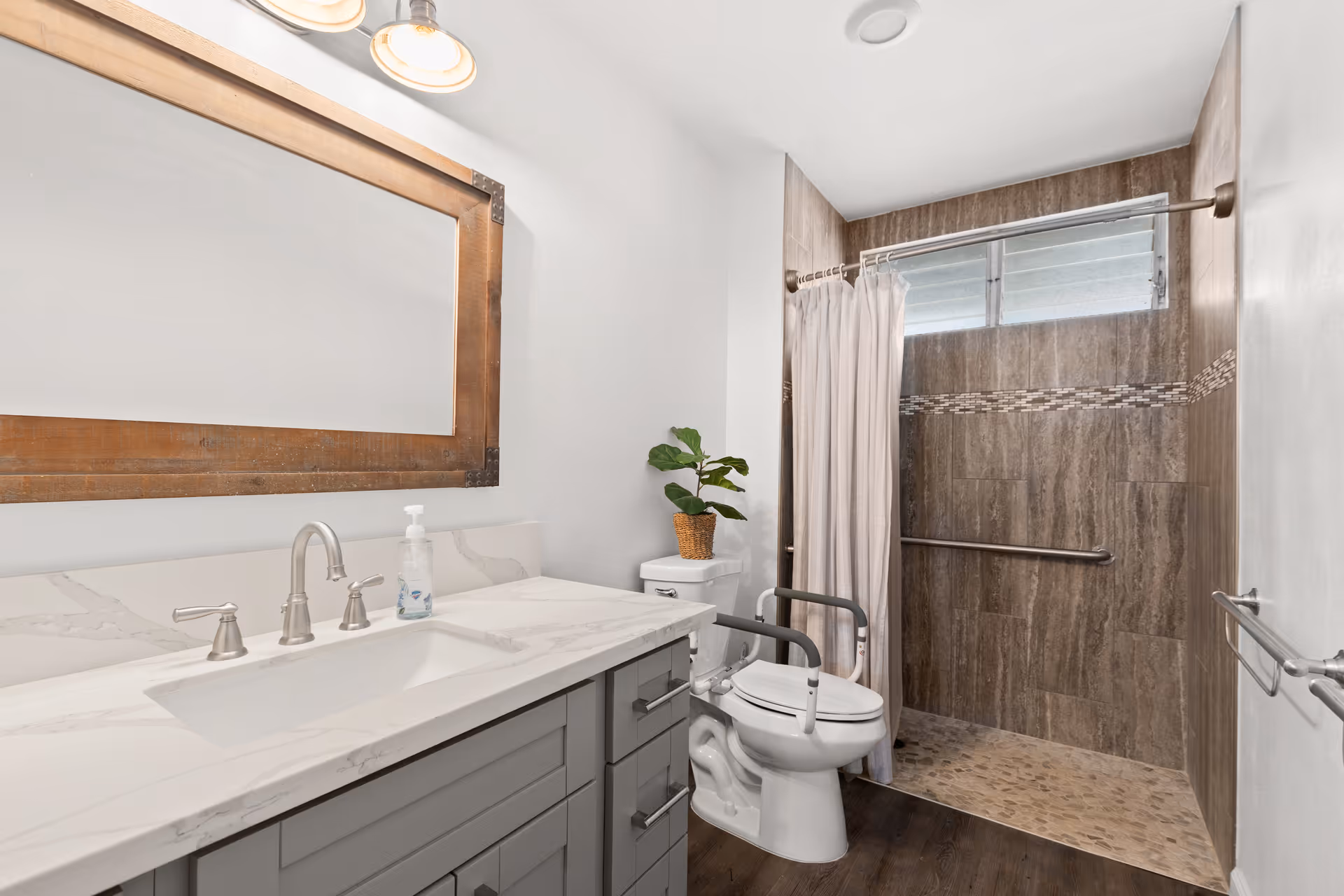 Bright accessible bathroom with a marble-top vanity and large mirror, a toilet with support rails, and a roll-in tiled shower with grab bars.