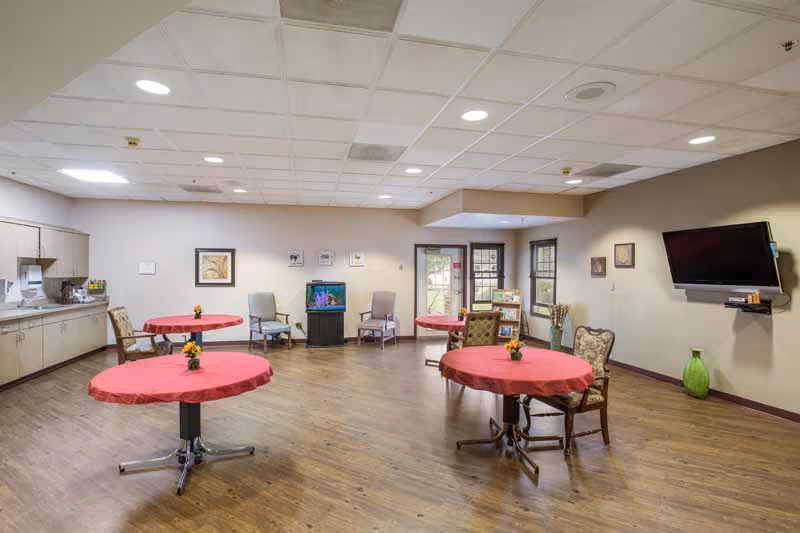 A spacious common room with wooden flooring and a drop ceiling. The room contains several round tables covered with red tablecloths, each with a small flower arrangement. There are various chairs around the tables and along the walls. A flat-screen TV is mounted on the right wall, and a small aquarium is positioned against the far wall. Cabinets and a countertop with a sink are on the left side of the room. Windows and a door provide natural light from the back wall.