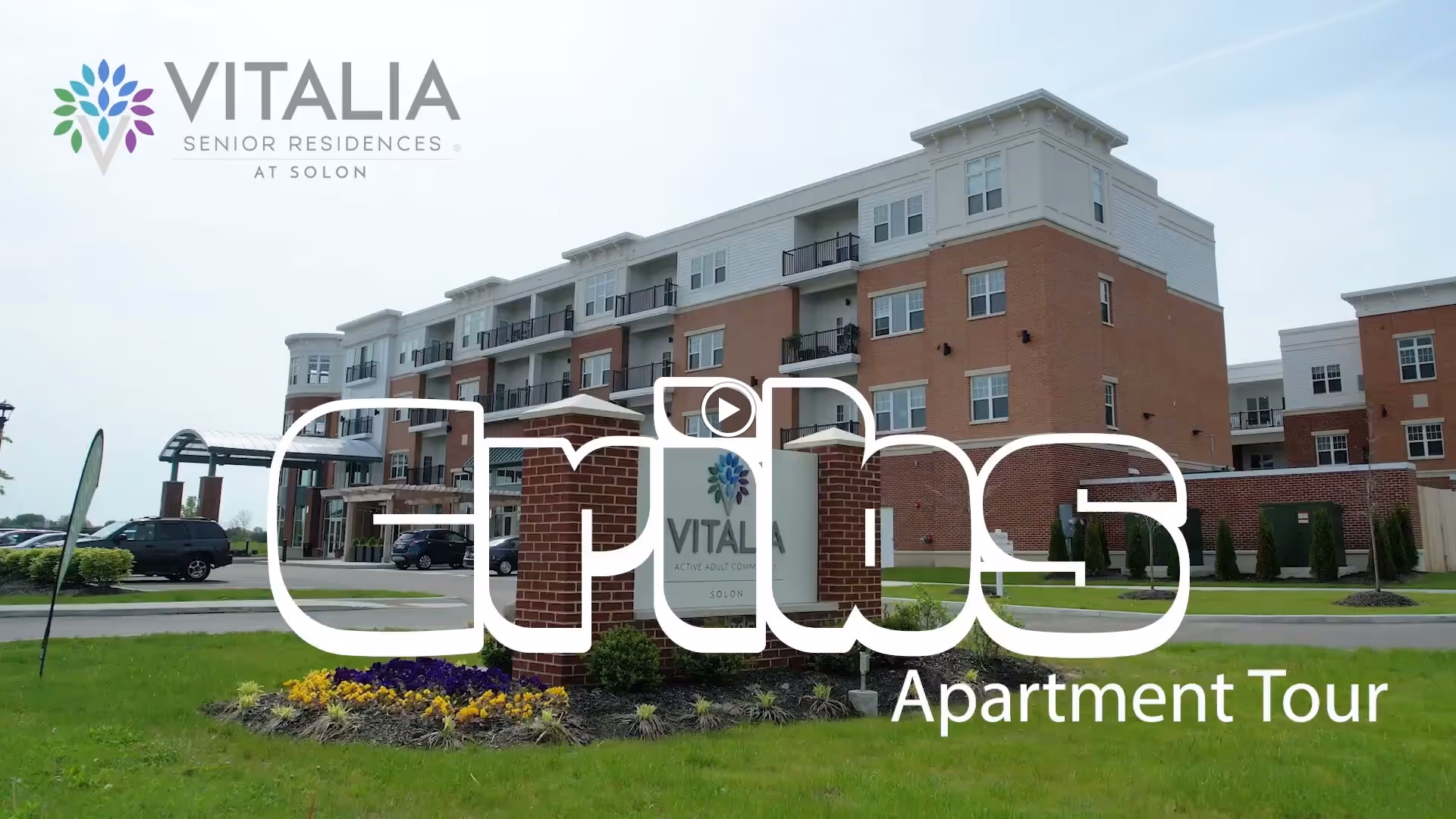 Exterior view of Vitalia Senior Residences at Solon, a multi-story brick and white building with balconies, surrounded by green grass and a flower bed in front of a sign displaying the community name.