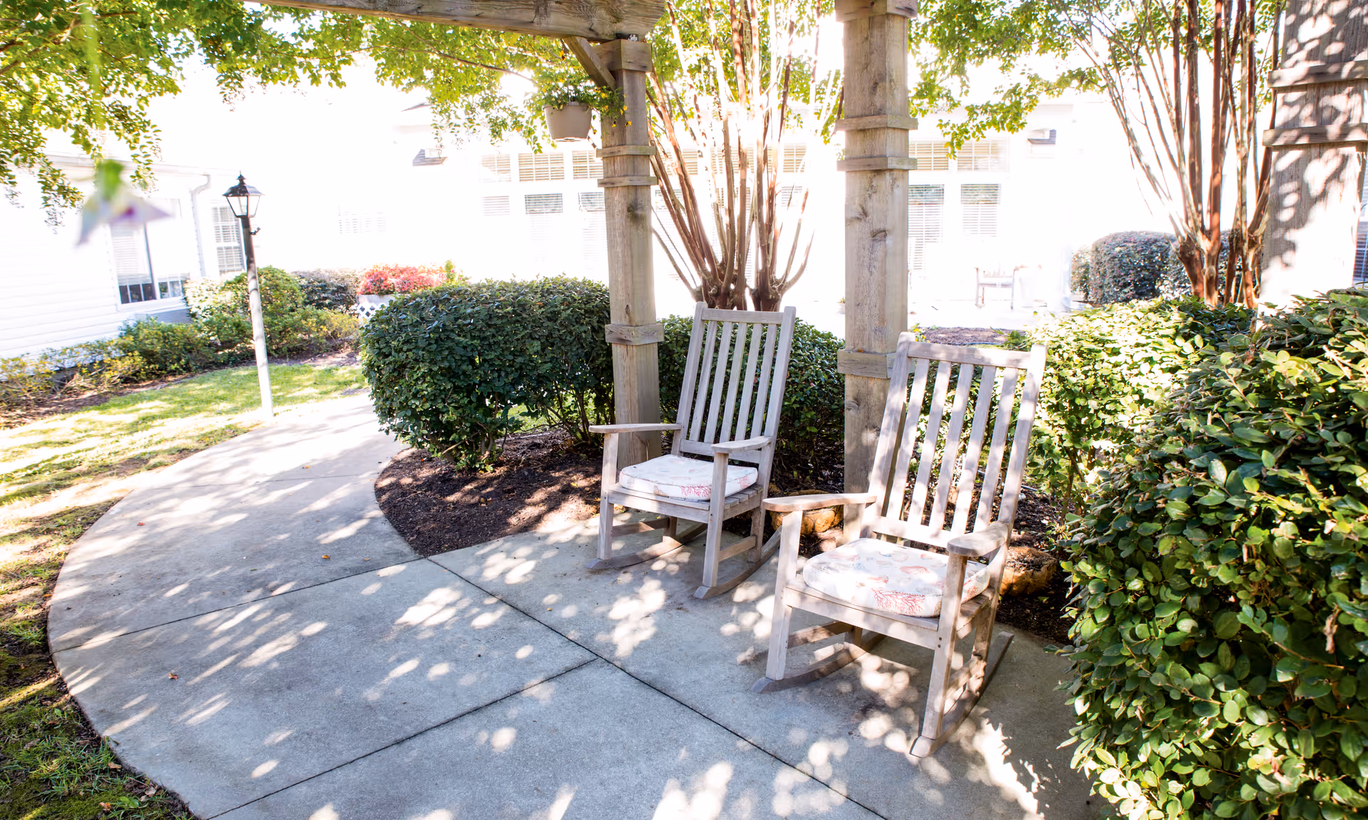 Two wooden rocking chairs with cushions sit under a pergola along a paved path in a sunlit garden courtyard.