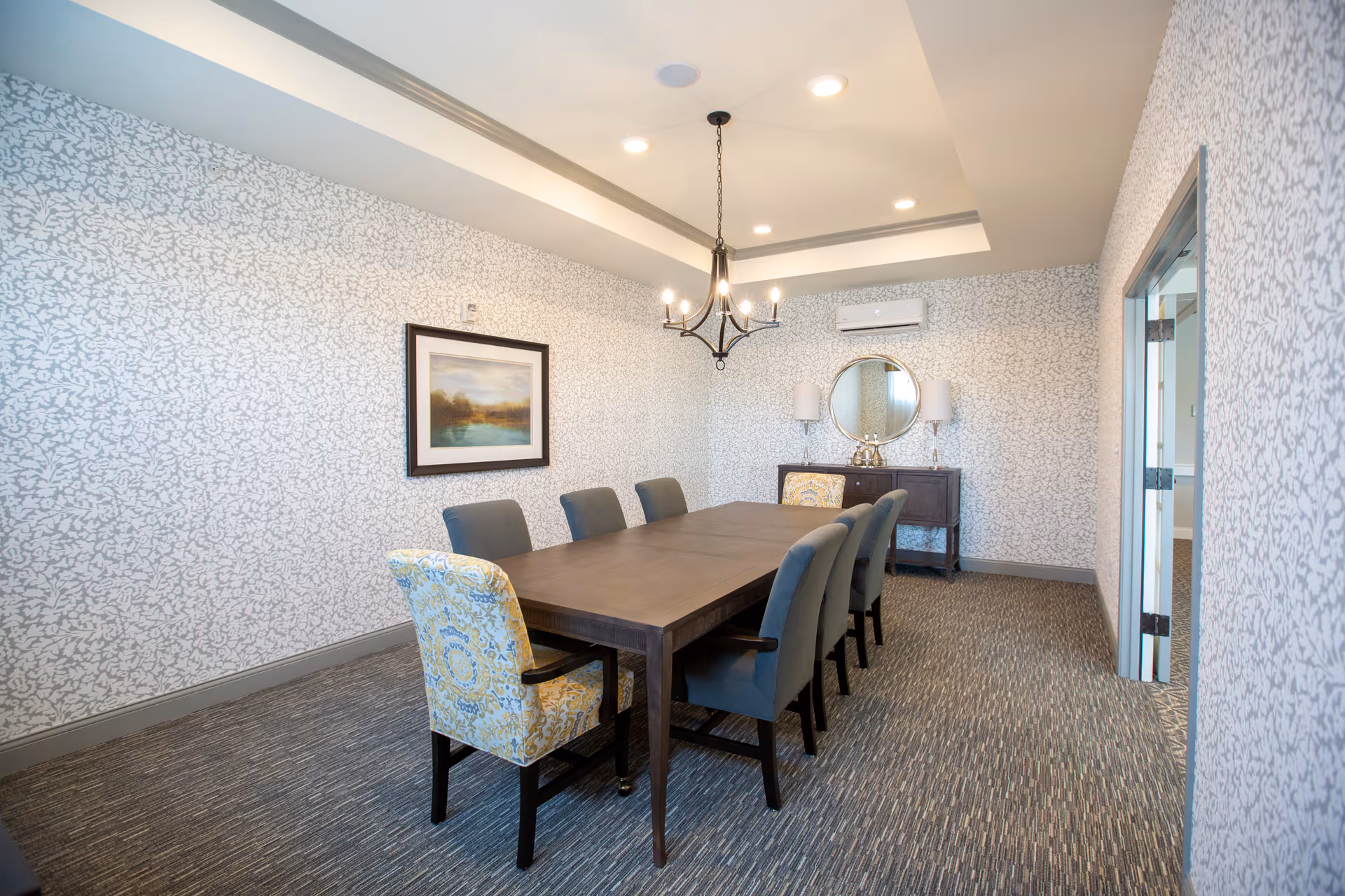 Long dining/meeting room with a wooden table surrounded by upholstered chairs, patterned wallpaper, chandelier, and a sideboard with a round mirror.