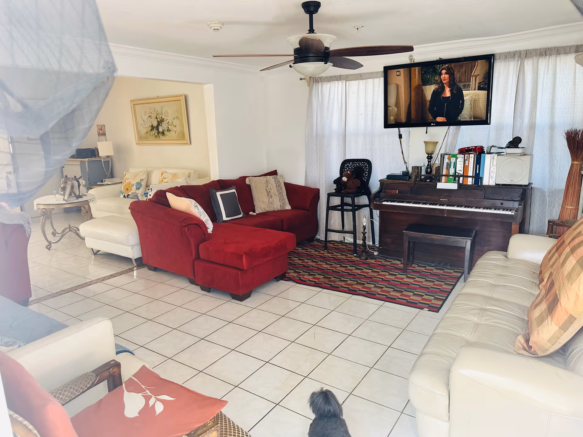 A bright living room with white tiled floors and white walls. The room features a red sectional sofa with multiple pillows, a white leather sofa with a plaid pillow, and a white armchair with a red cushion. A colorful patterned rug is placed in front of a dark wooden piano, which has books, binders, and a small lamp on top. A flat-screen TV is mounted on the wall above the piano, showing a woman speaking. There is a small black chair with a teddy bear on it next to the piano. In the background, there is a small table with a lamp and framed artwork on the wall.