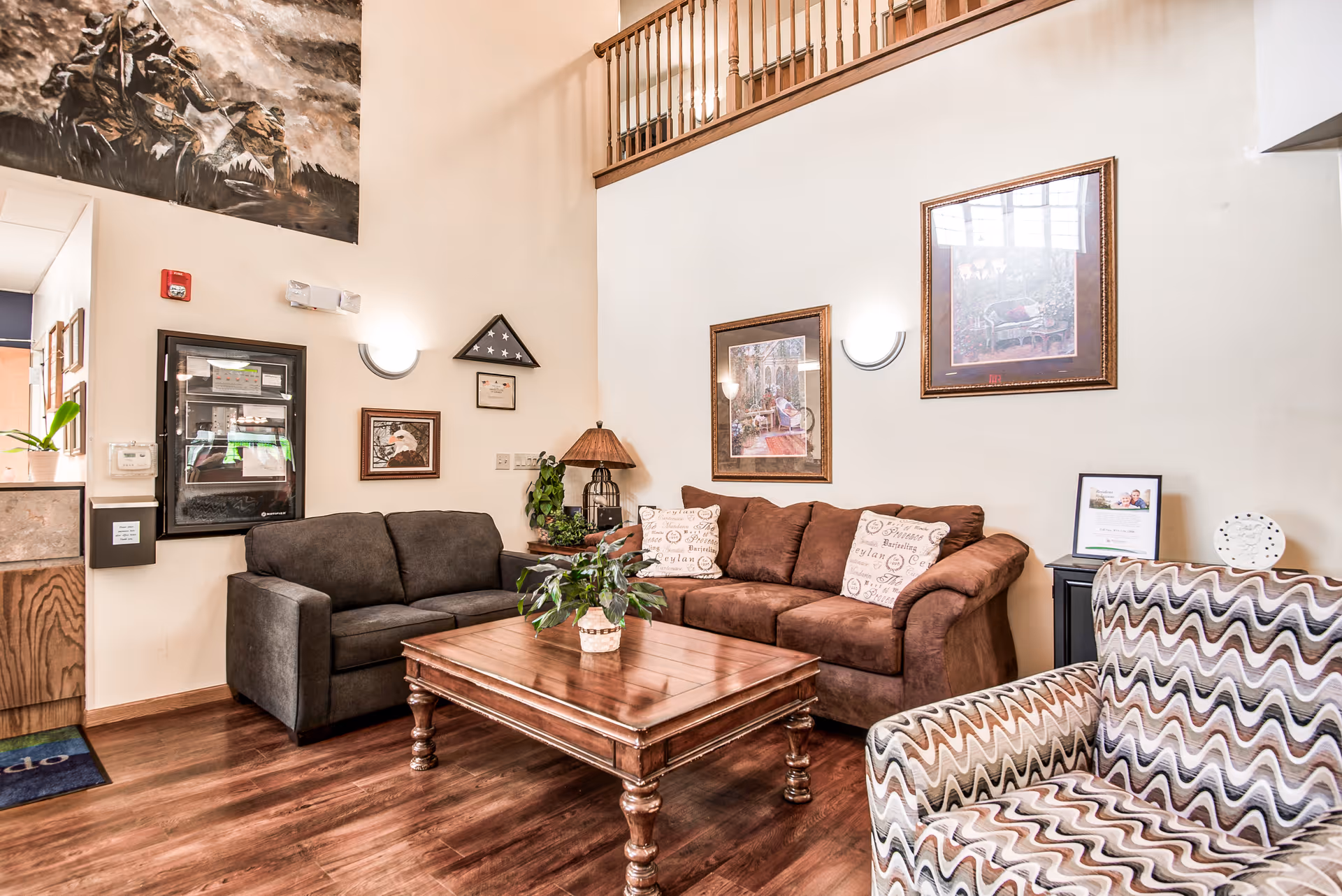 A cozy lounge with a brown sofa, patterned armchair, wooden coffee table, and framed artwork on a high wall.