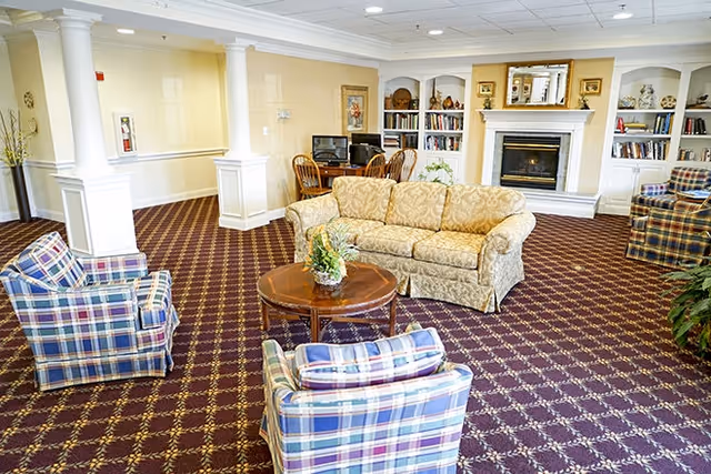 A cozy living room area in a senior living facility featuring a beige patterned sofa, two plaid armchairs, a round wooden coffee table with a floral arrangement, a fireplace with a mirror above it, built-in bookshelves filled with books and decorative items, and a small desk with two chairs and computers in the background.
