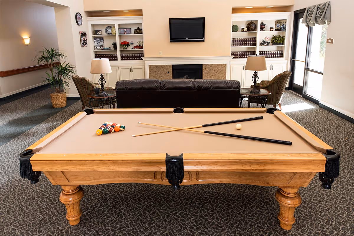 A cozy common area with a pool table in the foreground, two pool cues crossed on the table, a set of billiard balls arranged for a game, a brown leather couch, two armchairs with side tables and lamps, built-in shelves with decorative items and books, a wall-mounted TV above a fireplace, and a large window with drapes letting in natural light.