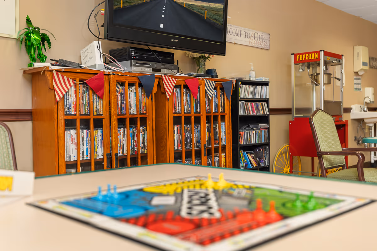 A cozy common area with a board game on a table in the foreground, a wooden cabinet filled with DVDs and books, a television mounted on the wall, a popcorn machine, and a small bookshelf with CDs. There are chairs around the table and a small sink in the background.