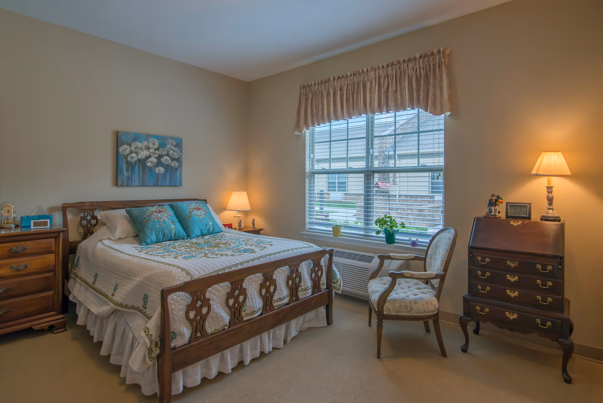 A cozy, traditionally furnished bedroom with a wooden bed, bedside tables, an upholstered chair, and a large window letting in daylight.