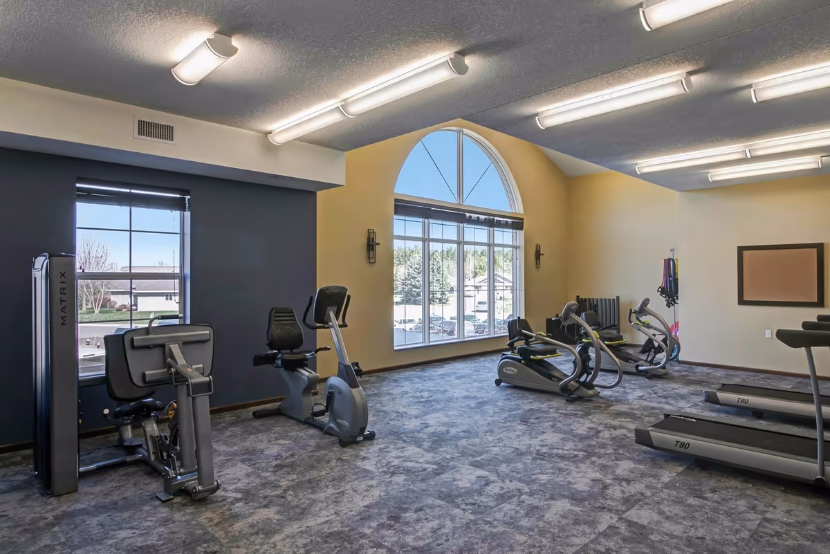 A well-lit fitness room with exercise equipment including treadmills, stationary bikes, and strength training machines. The room has large windows with a view of trees and a parking lot outside, and the walls are painted in neutral tones with a high ceiling featuring fluorescent lights.