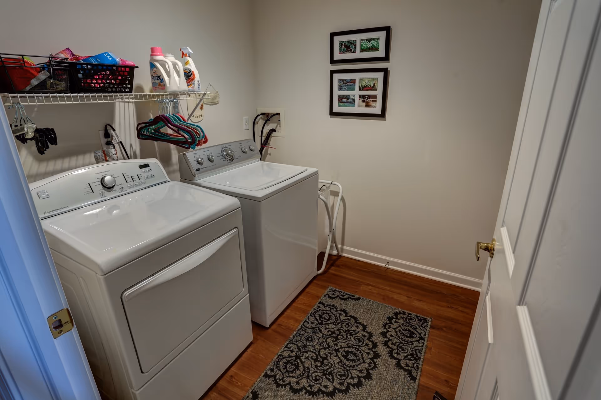 Laundry room with a white washing machine and dryer side by side, a shelf above holding laundry detergent and hangers, a patterned rug on the wooden floor, and two framed pictures on the wall.