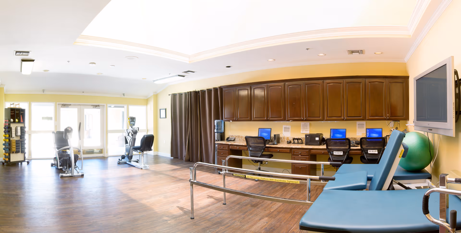 A spacious rehabilitation room with exercise equipment including stationary bikes, a blue therapy table, a large green exercise ball, and a wall-mounted TV. There are three computer workstations with chairs along a wall with wooden cabinets above. The room has wooden flooring and large windows letting in natural light.