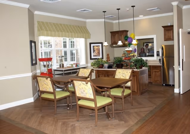 Interior dining area with a wooden table and four floral-upholstered chairs, a kitchen island with plants and pendant lights, refrigerator, and a large window.