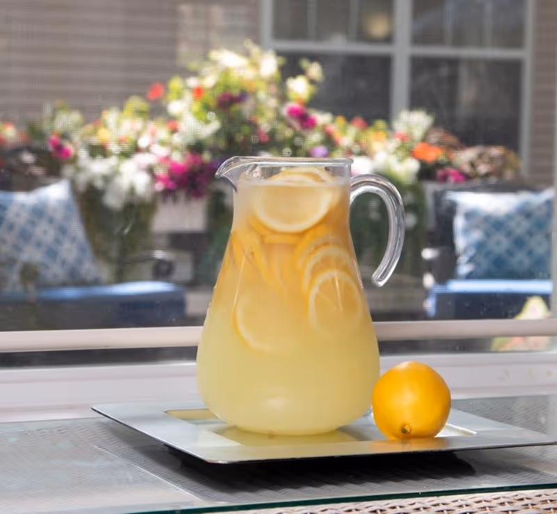 A glass pitcher filled with lemonade and lemon slices placed on a tray next to a whole lemon, with a blurred background showing outdoor seating and colorful flowers.