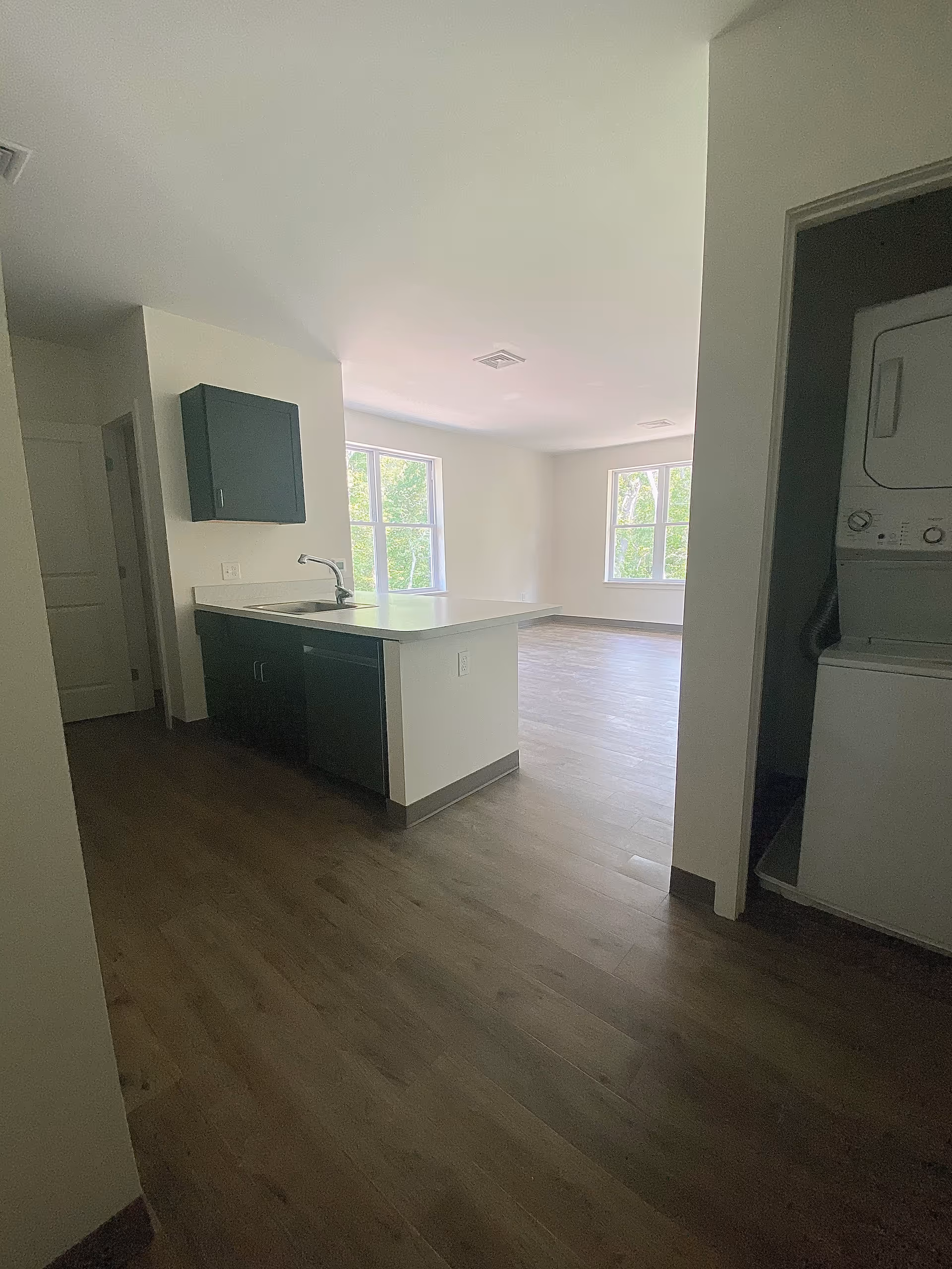 Interior view of an apartment with a kitchen area featuring a sink and cabinets, a laundry closet with a stacked washer and dryer, and a bright living area with large windows letting in natural light.