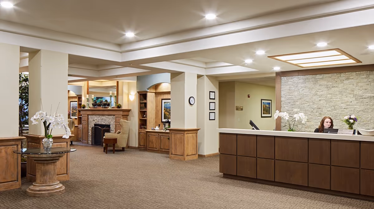 Reception area of a senior living facility with a front desk where a woman is seated. The space features warm lighting, wooden cabinetry, a stone accent wall behind the desk, a fireplace with seating in the background, and decorative plants including white orchids.