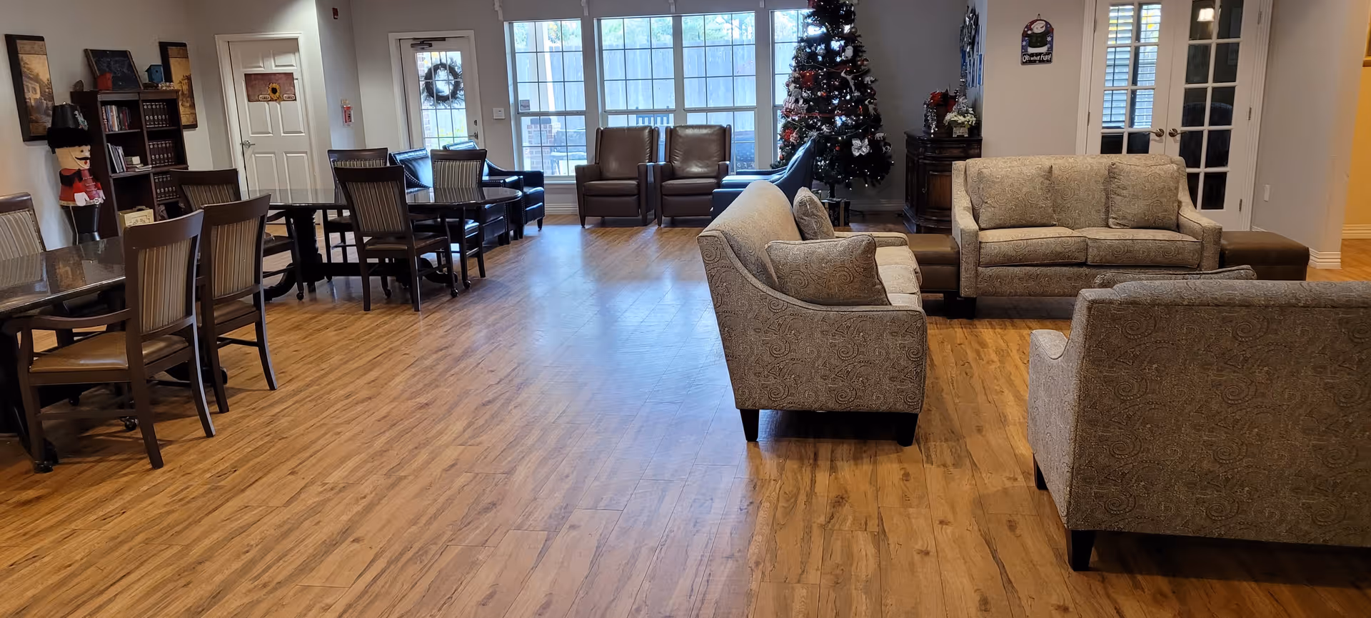 A spacious senior living common area with wooden flooring, featuring multiple seating arrangements including sofas, armchairs, and dining tables with chairs. A decorated Christmas tree is visible near large windows letting in natural light. The room has a warm and inviting atmosphere with neutral-colored walls and furniture.