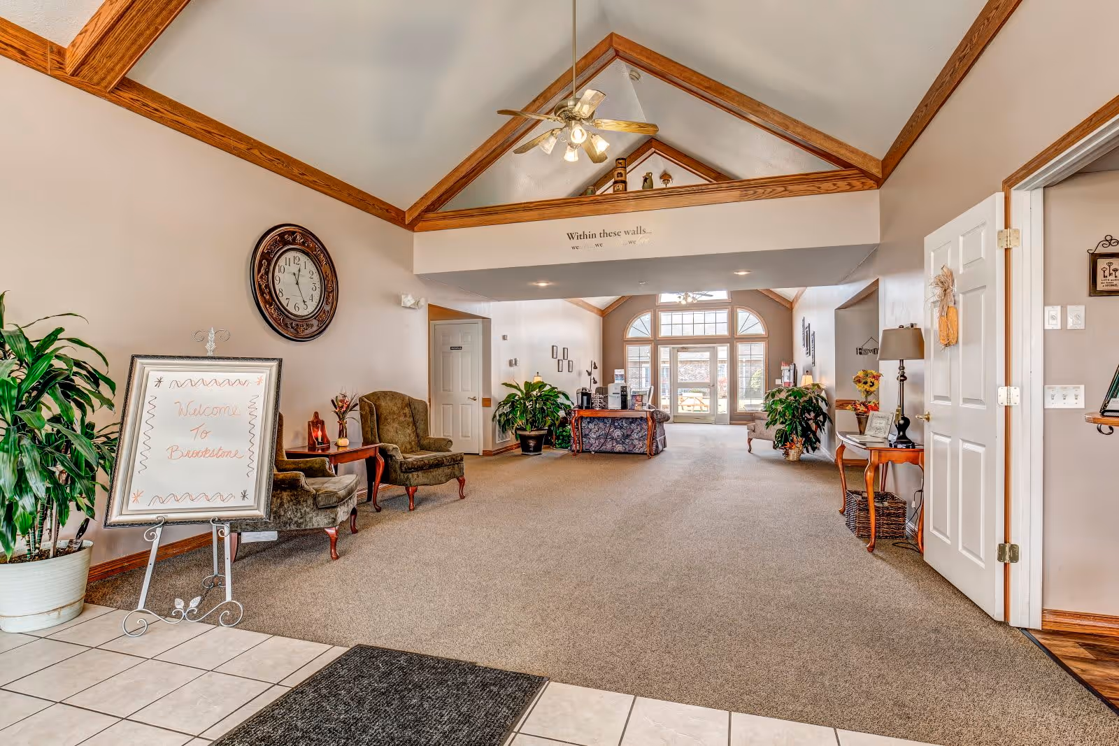 Spacious and well-lit lobby area of Brookstone Estates Of Fairfield with high vaulted ceilings and wooden beams. The room features a large clock on the wall, two upholstered armchairs with a small table between them, potted plants, a welcome sign on an easel, and a reception desk in the background near large windows letting in natural light.