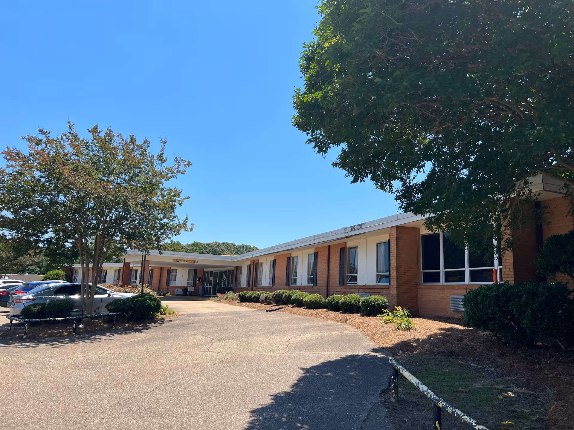 Front exterior of a one-story brick retirement center with a driveway, parked cars, and trees.