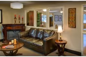 A cozy living room area featuring a dark brown leather sofa, a round wooden coffee table with books and candles, a wooden side table with a lamp, and a decorative fireplace with candles on the mantel. The walls are adorned with framed artwork and mirrors, creating a warm and inviting atmosphere.