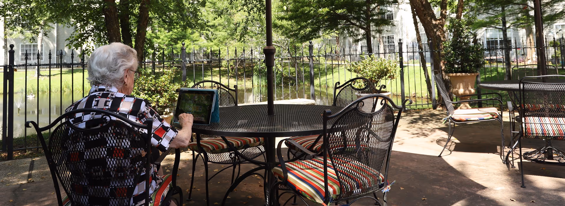 An elderly woman with white hair sitting outdoors at a metal table with striped cushions, using a tablet. The area is shaded by trees and surrounded by a black metal fence, with a pond and greenery visible in the background.