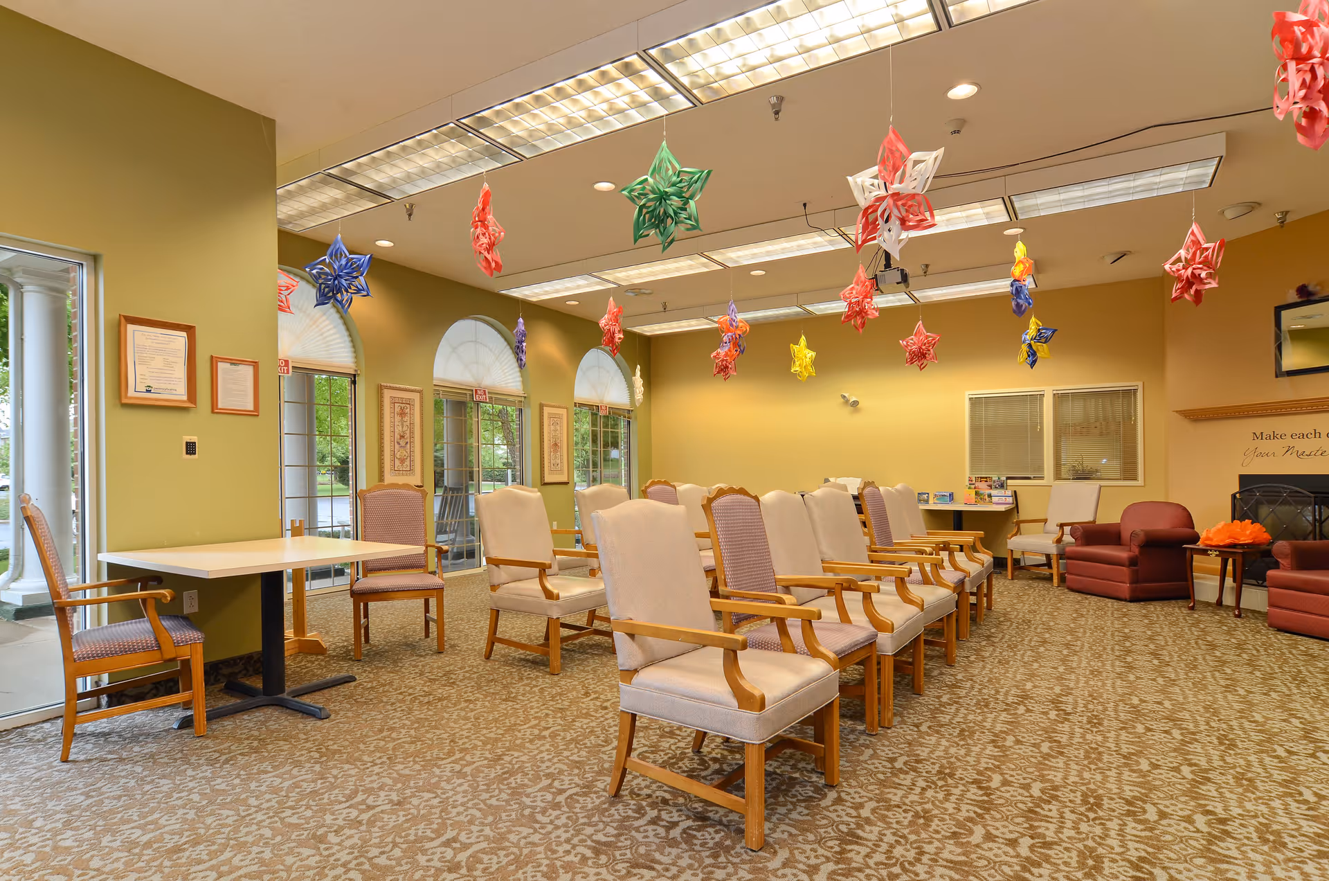 A spacious room with rows of beige cushioned chairs with wooden armrests arranged facing forward. The ceiling has fluorescent lights and colorful star-shaped paper decorations hanging down. The walls are painted light yellow and green, with large arched windows letting in natural light. There are a few armchairs and small tables along the walls, and framed certificates and artwork are displayed. The room has a patterned carpet and appears to be a common area or meeting room in a senior living facility.