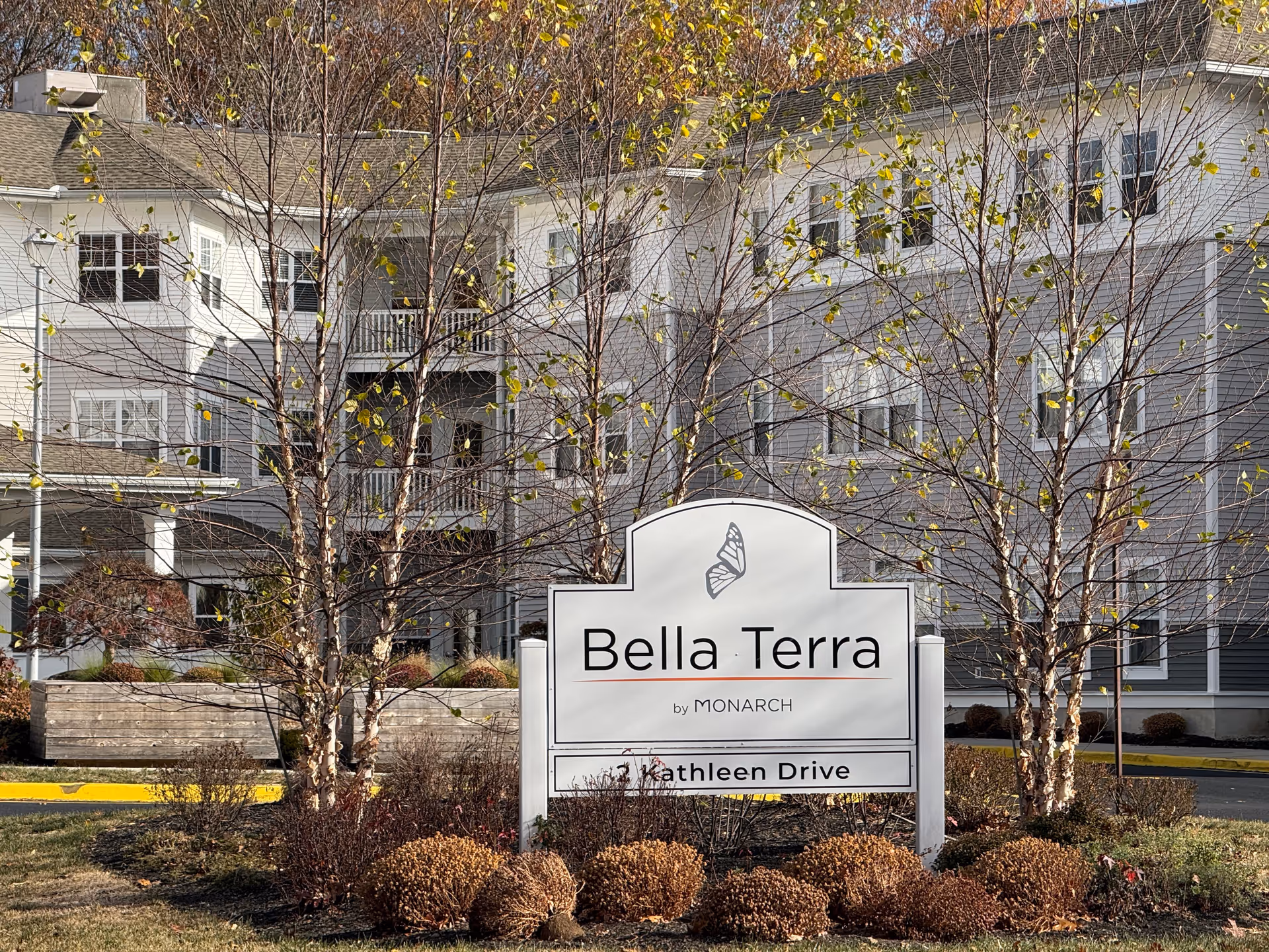 Outdoor view of the Monarch Bella Terra senior living facility sign with a butterfly logo, surrounded by trees and shrubs, with the building visible in the background.