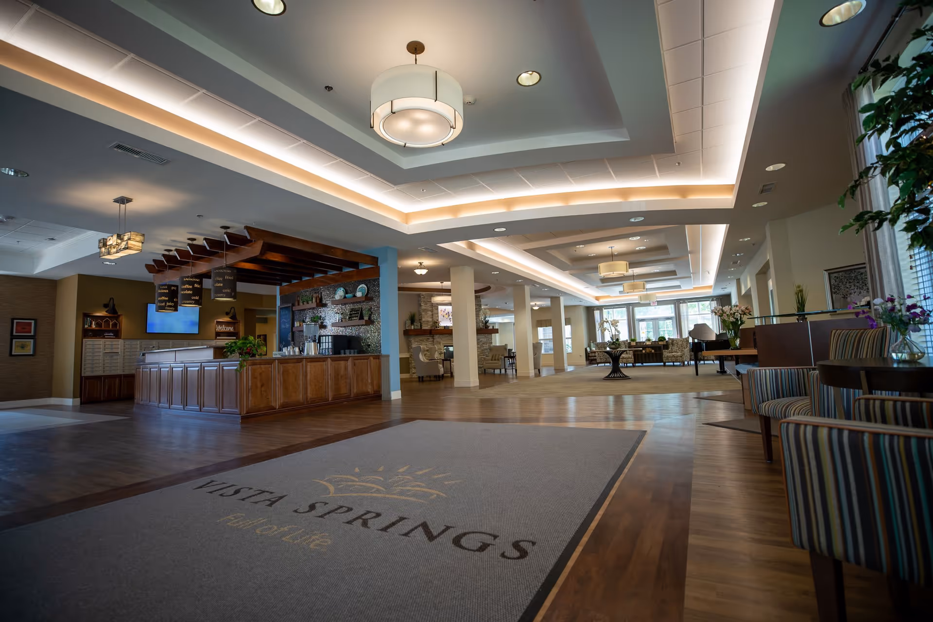 Spacious, well-lit senior living lobby with a reception desk, seating areas, and a floor mat reading 'Vista Springs'.