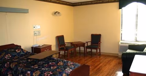 A furnished senior living bedroom with a bed, overbed table, two chairs and small table, a couch, and a window with blinds.