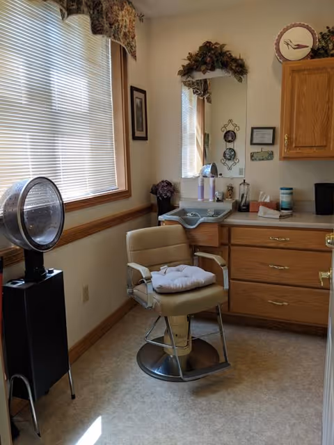A small room with a beige salon chair with a cushion on it, a hair dryer on a stand, a countertop with various items including a tissue box and bottles, a mirror with floral decoration above it, and a window with blinds and a floral valance.