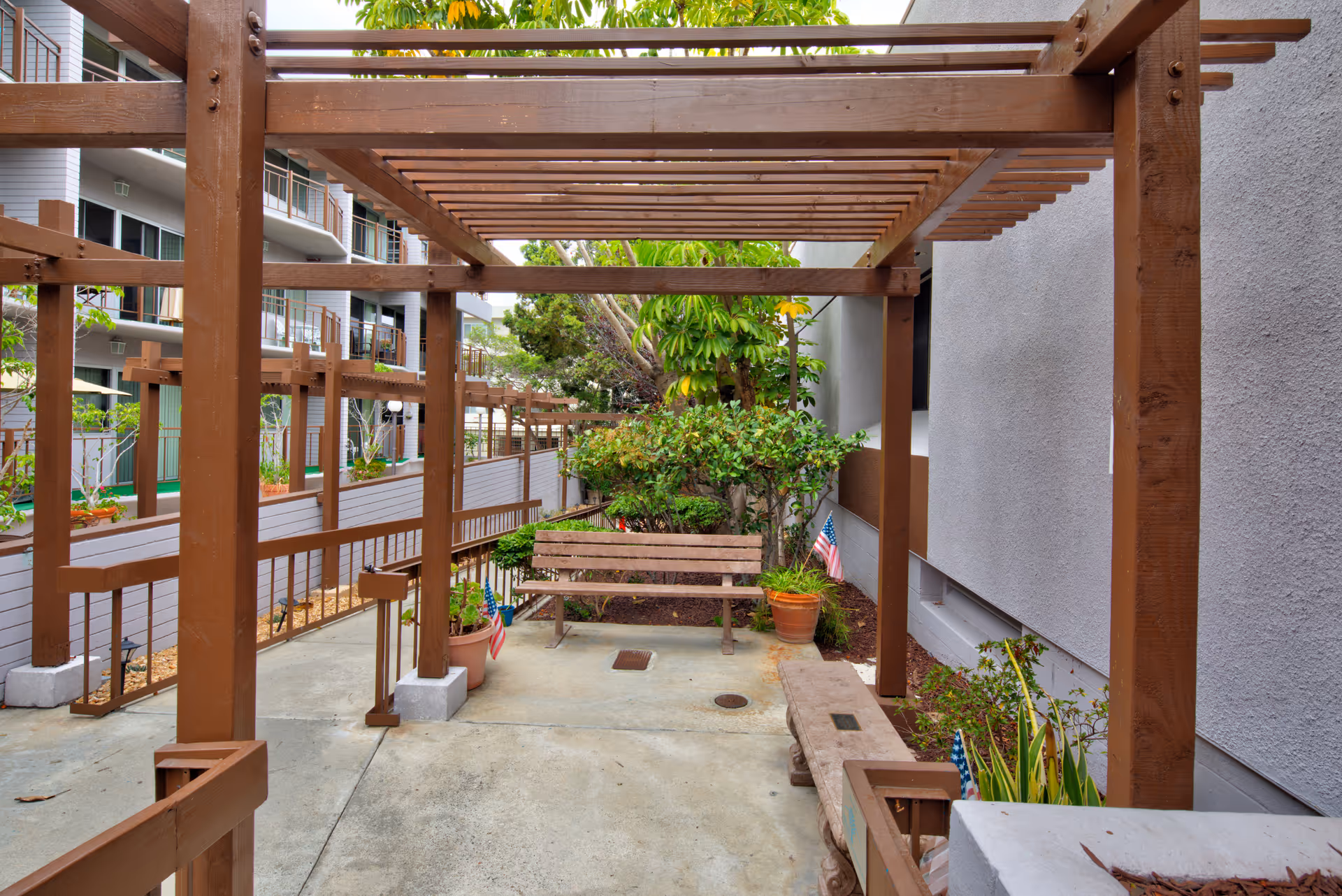 Outdoor seating area at a senior living facility featuring wooden pergolas, benches, potted plants, and small American flags, with a multi-story building visible in the background.