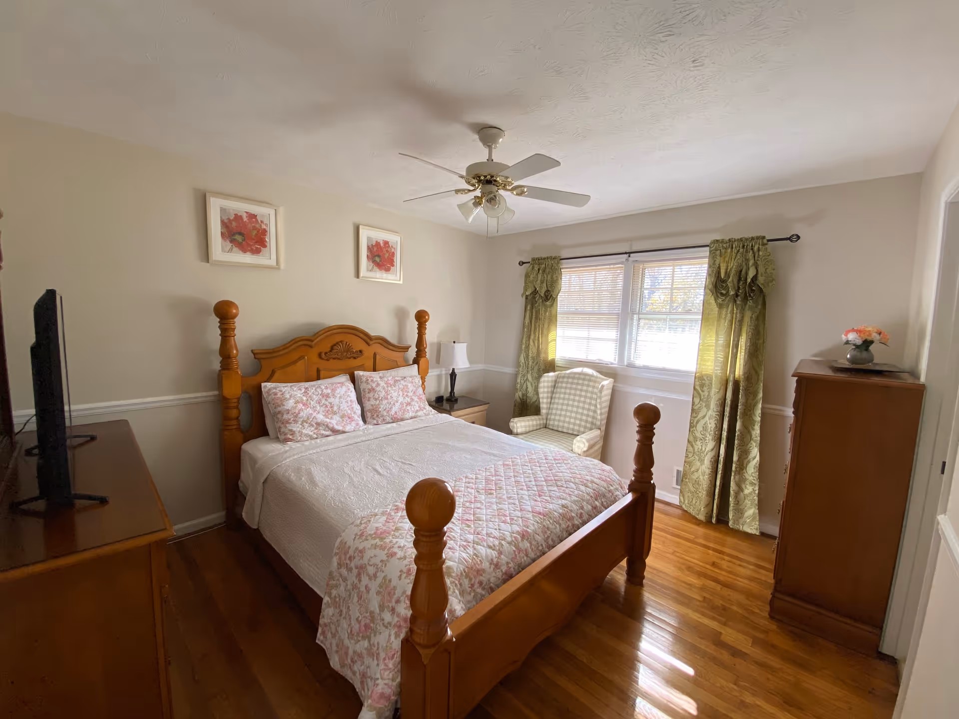 A cozy bedroom with a wooden bed frame, white and pink floral bedding, two framed flower pictures on the wall, a ceiling fan with lights, a window with green curtains, a checkered armchair, a wooden dresser with a vase of flowers, and a television on a wooden stand.