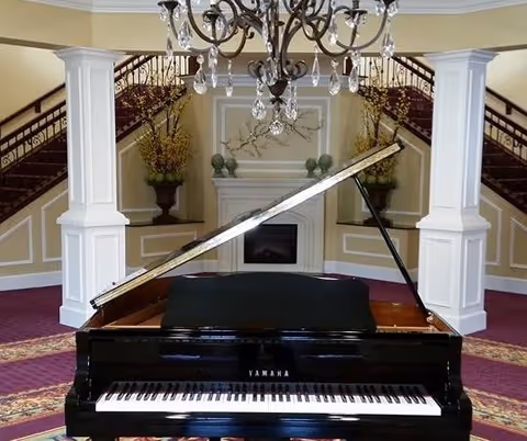 A grand piano with its lid open is positioned in the center of a room with a purple carpet and patterned rug. Behind the piano, there is a fireplace flanked by two white columns and decorative plants. A chandelier hangs from the ceiling, and a staircase with wooden railings curves upward on both sides of the fireplace.