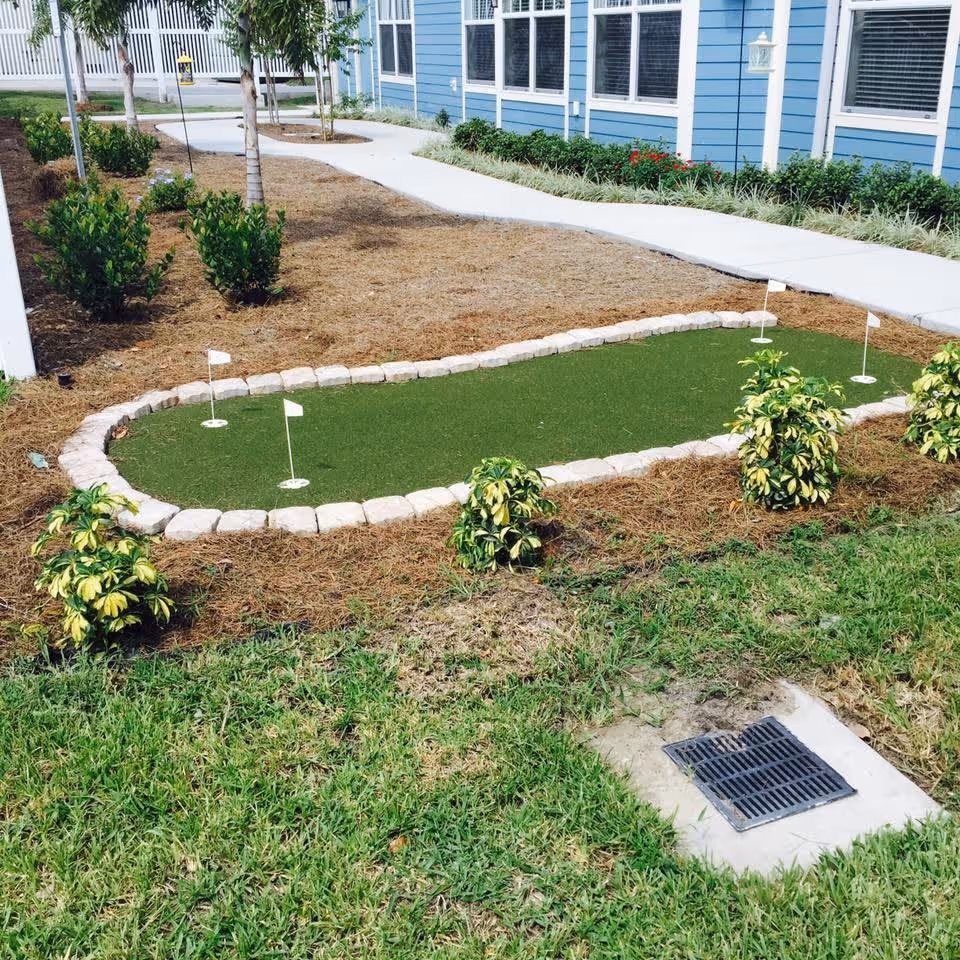 A small outdoor putting green with four holes and white flags, surrounded by stone edging and small bushes. A concrete walkway runs alongside a blue building with white trim and several windows. There are trees and landscaped areas with mulch around the putting green.