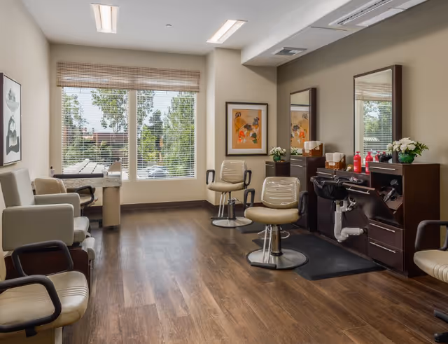 Salon-style common room with styling chairs, mirrors, a shampoo sink, and large windows.