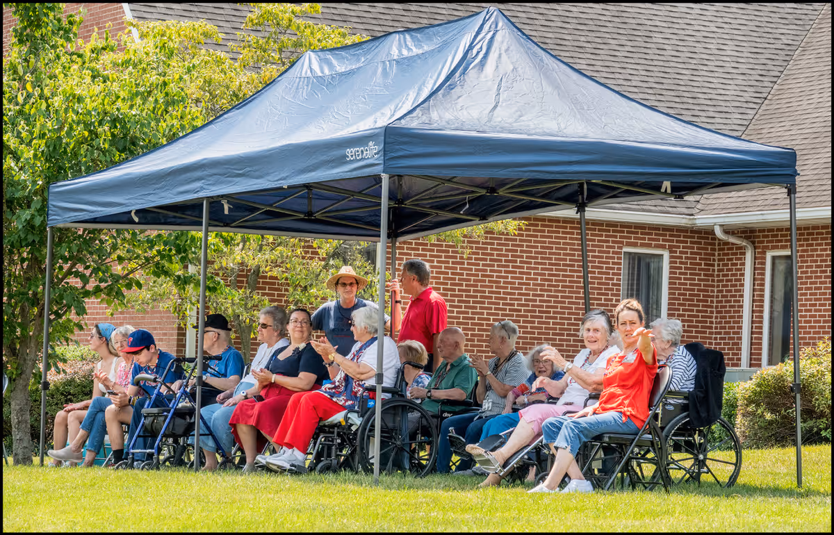 A group of elderly people sitting under a blue canopy tent on a grassy area outside a brick building. Some are in wheelchairs, and a few caregivers or visitors are standing or sitting with them. The scene is bright and sunny with green trees in the background.