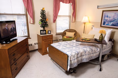 A cozy bedroom in a senior living facility featuring a single bed with a patterned quilt, a wooden dresser with a television on top, a small wooden cabinet with a small decorated Christmas tree and framed photos, a wicker chair with cushions, and a floor lamp. The room has two windows with pink curtains and a framed painting on the wall.