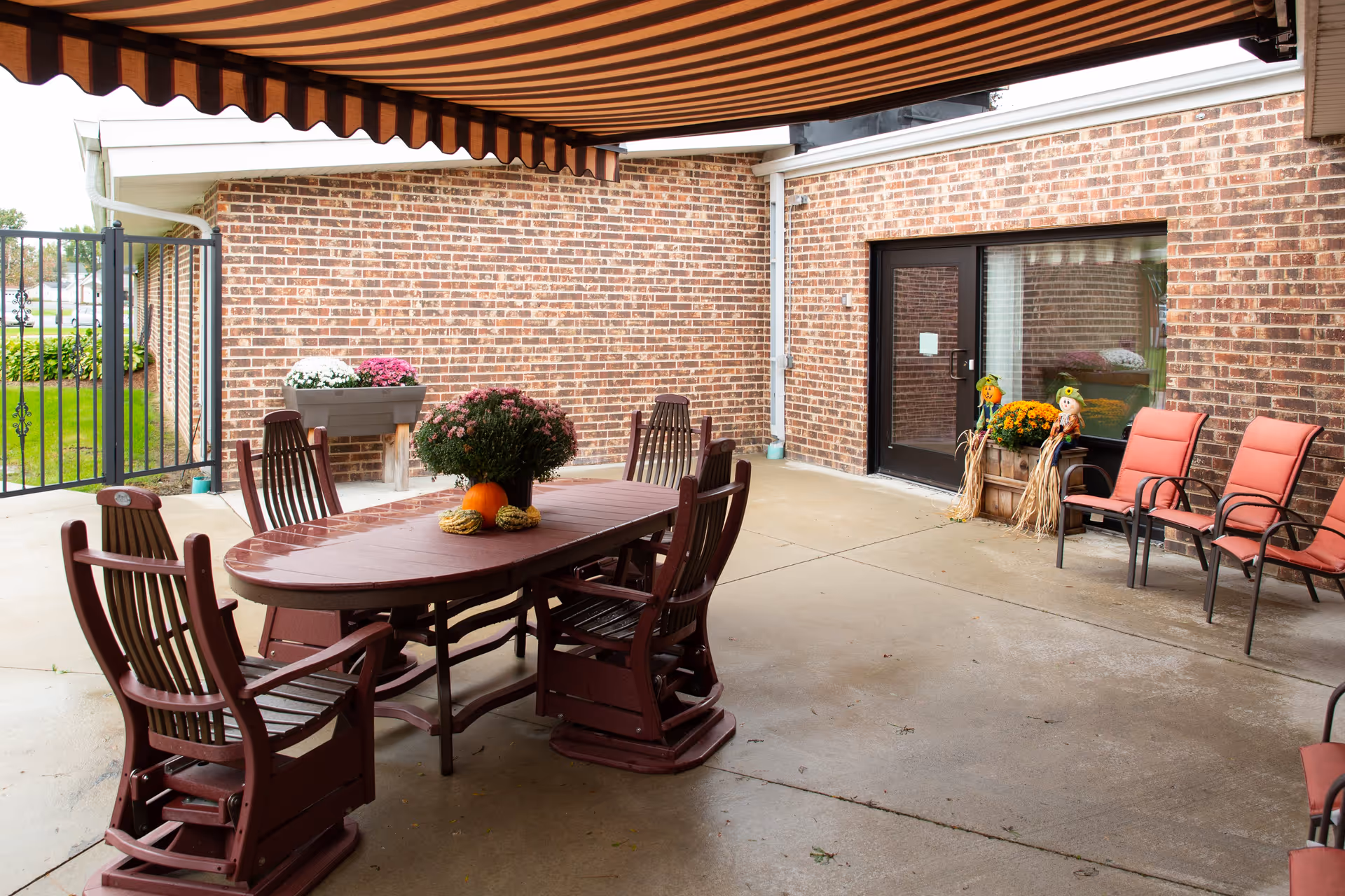 Covered outdoor patio area with a wooden table and six wooden chairs. The table has a centerpiece with flowers and small pumpkins. There are additional chairs with red cushions along the brick wall, which has a door and a window. Decorative scarecrows and flower pots are placed near the door. A striped awning provides shade over the patio.