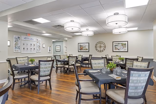 A dining room with multiple tables covered in dark tablecloths, each set with glasses, napkins, and silverware. The room has wooden flooring, cushioned chairs with patterned backs, and modern ceiling lights. The walls are decorated with framed pictures and a large clock.