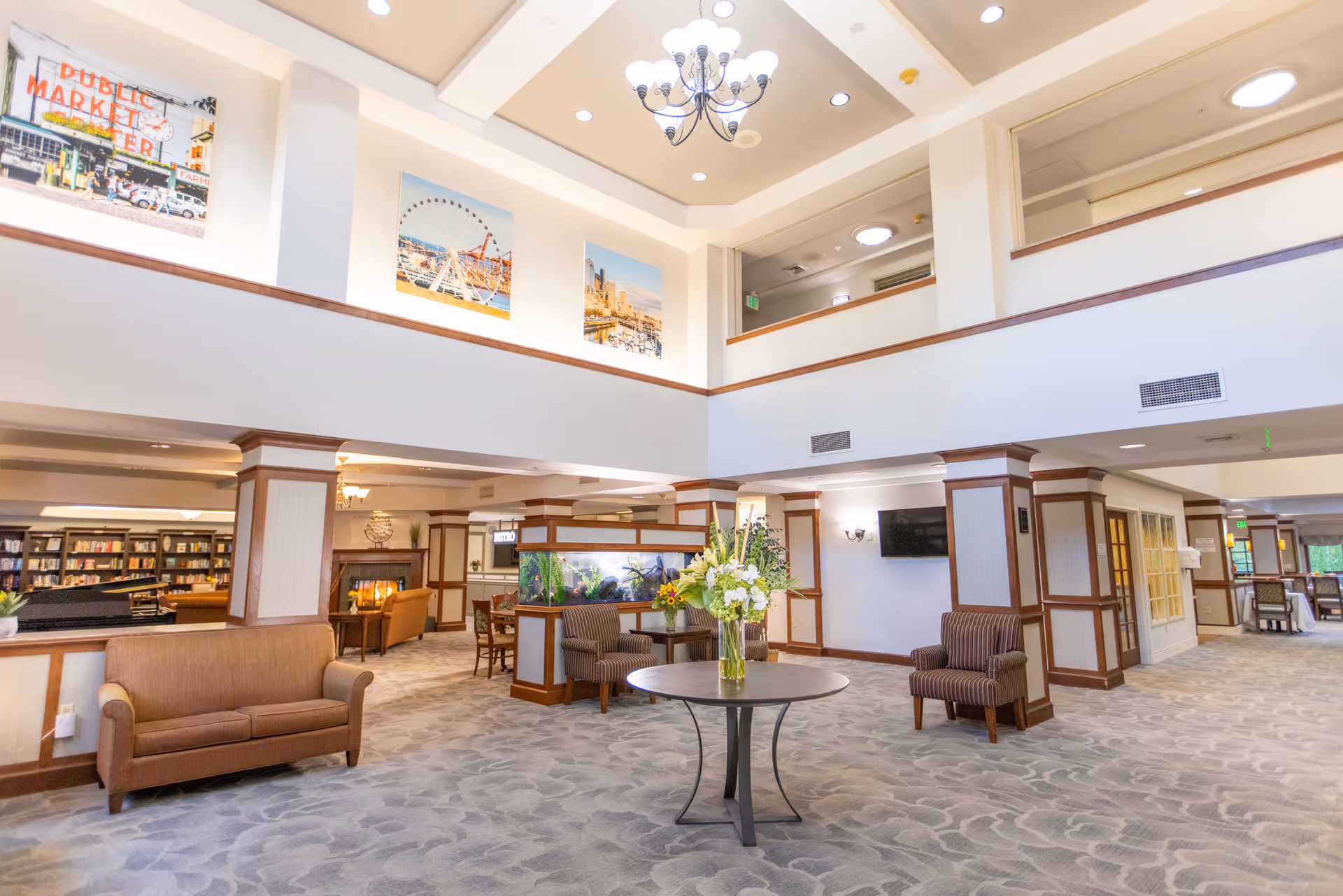 Spacious and well-lit senior living facility common area with high ceilings, a chandelier, comfortable seating including sofas and armchairs, a round table with a flower arrangement, an aquarium, and framed pictures on the walls. There is a fireplace and a bookshelf visible in the background.
