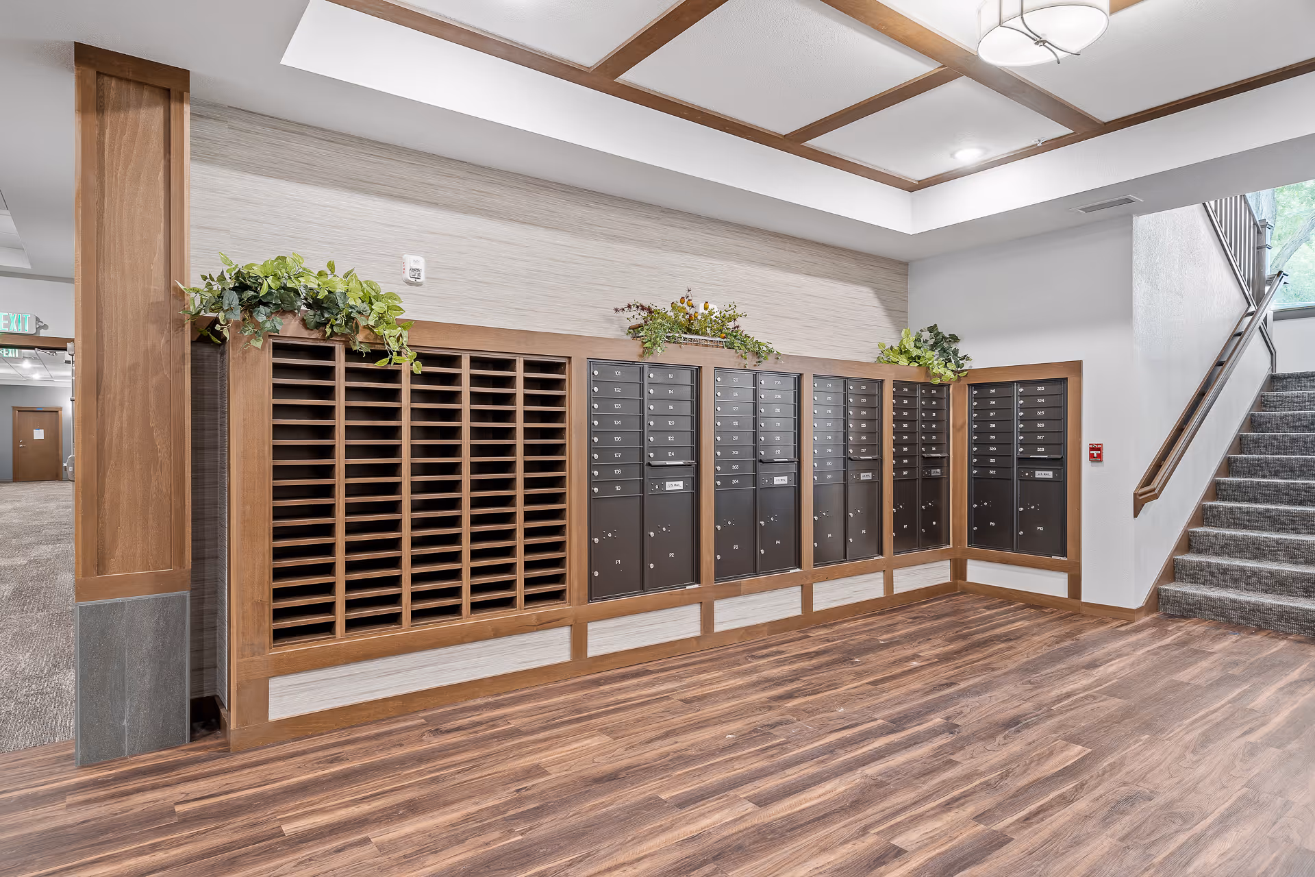 Interior view of a senior living facility mailroom area with multiple black mailboxes mounted on a wooden frame along the wall. Above the mailboxes are decorative green plants. To the right, there is a carpeted staircase with a wooden handrail leading upwards. The floor is wood-patterned, and the ceiling has recessed lighting with wooden beams.