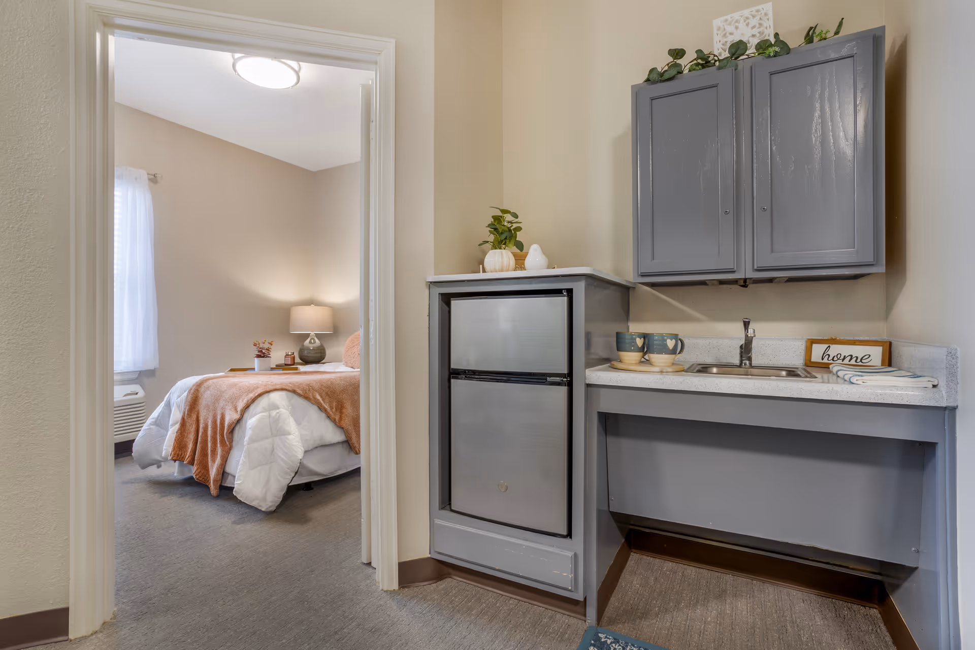 View of a small kitchenette area with gray cabinets, a mini refrigerator, a sink, and a countertop with two mugs and a small decorative sign that says 'home'. Through an open doorway, a bedroom is visible with a bed covered in white and orange bedding, a bedside table with a lamp, and a window with sheer curtains.