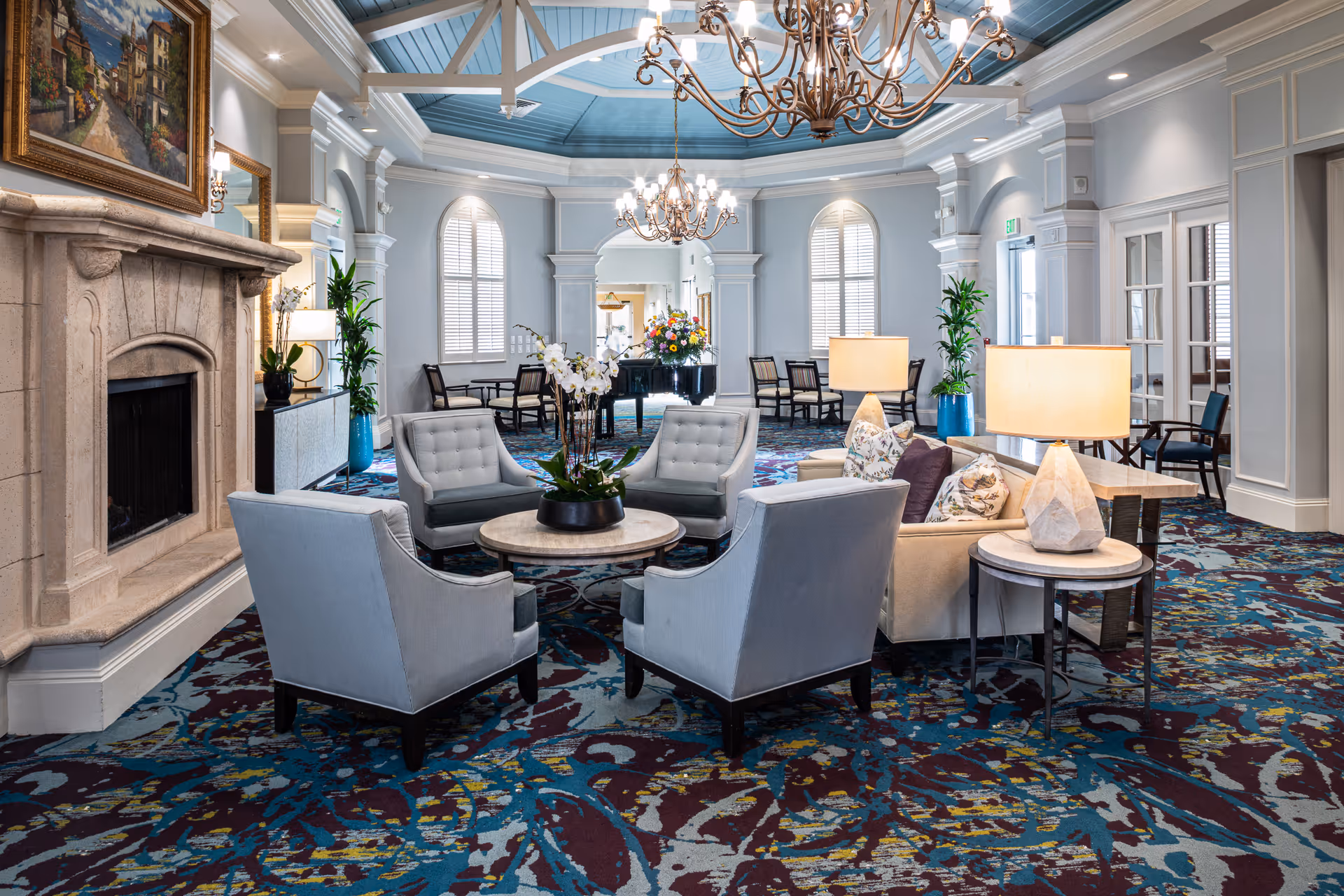 A spacious and elegant living room area with a patterned carpet, four gray upholstered armchairs arranged around a round coffee table with a potted orchid, a beige sofa with floral pillows, two table lamps, a stone fireplace with a painting above it, large windows with white shutters, and two ornate chandeliers hanging from a blue ceiling.
