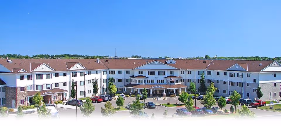 Front view of a large three-story assisted living building with a central covered entrance, landscaped grounds, and parked cars under a clear blue sky.