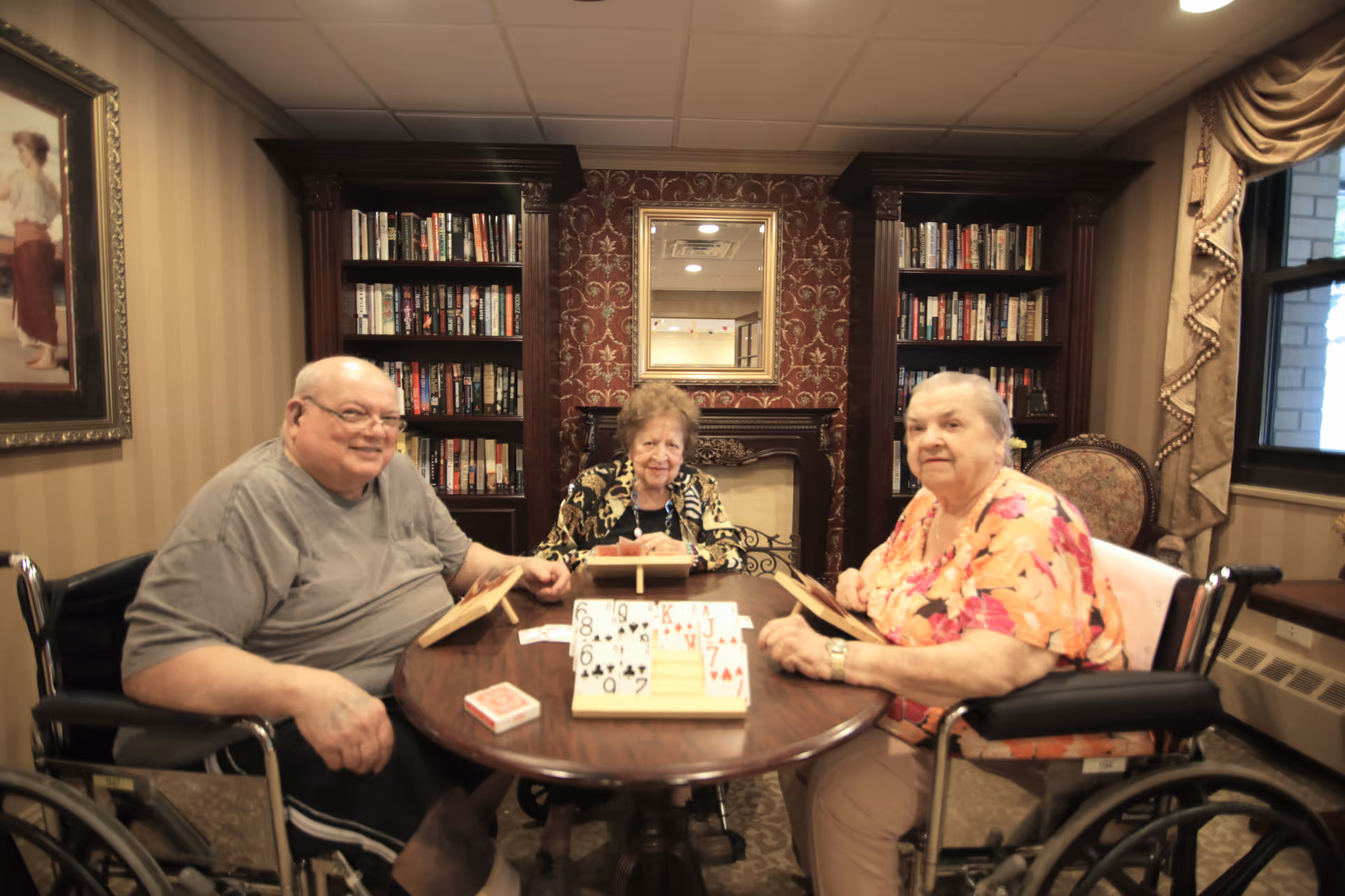 Three elderly individuals sitting around a round wooden table playing a card game in a cozy room with bookshelves, a fireplace, and a window with drapes. Two of the individuals are in wheelchairs, and the room has warm lighting and traditional decor.