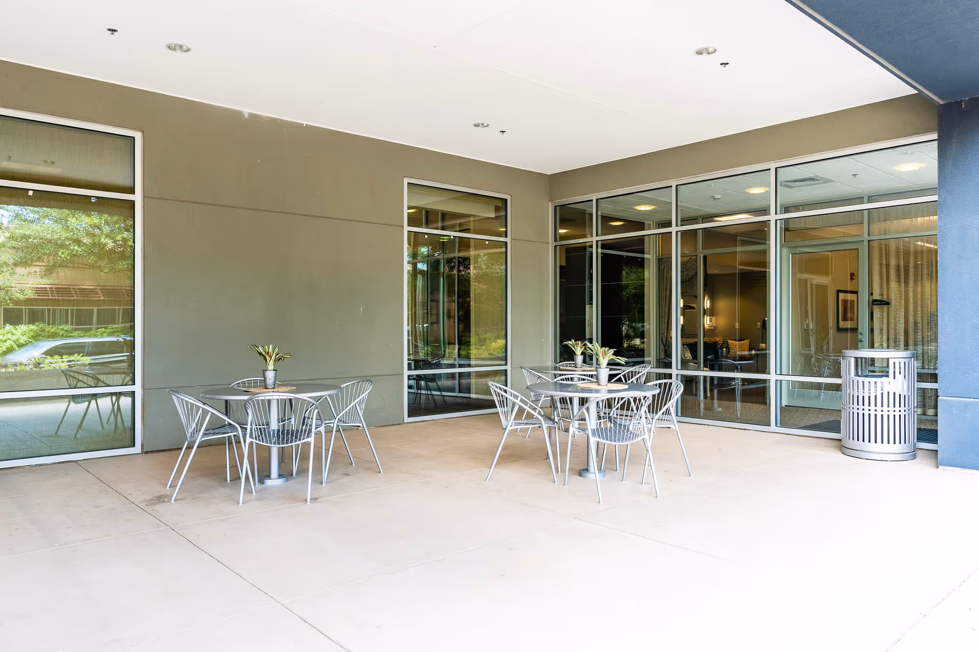 Covered outdoor patio with metal tables and chairs and potted plants in front of large glass windows.