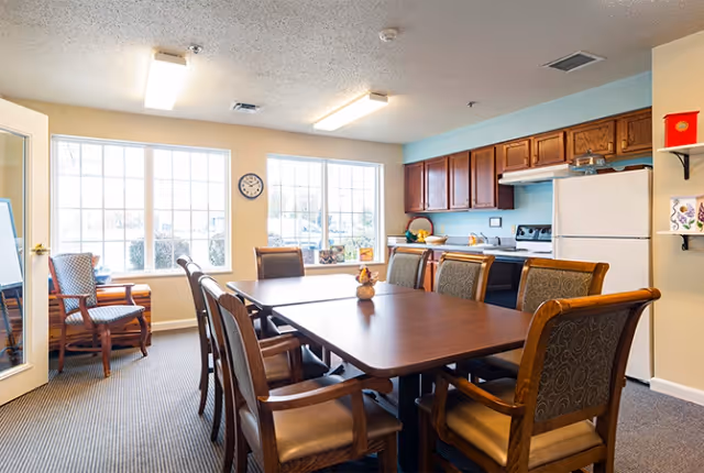 A bright dining area with a large wooden table surrounded by eight cushioned chairs. The room has two large windows letting in natural light, a clock on the wall, and a kitchen area with wooden cabinets, a white refrigerator, and a stove in the background. There are two additional chairs near the windows and a small decorative item on the table.