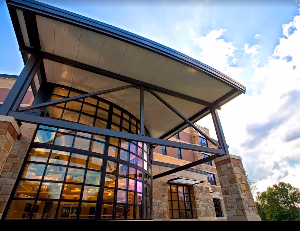 Modern building entrance with a large curved glass façade and metal canopy under a partly cloudy sky.