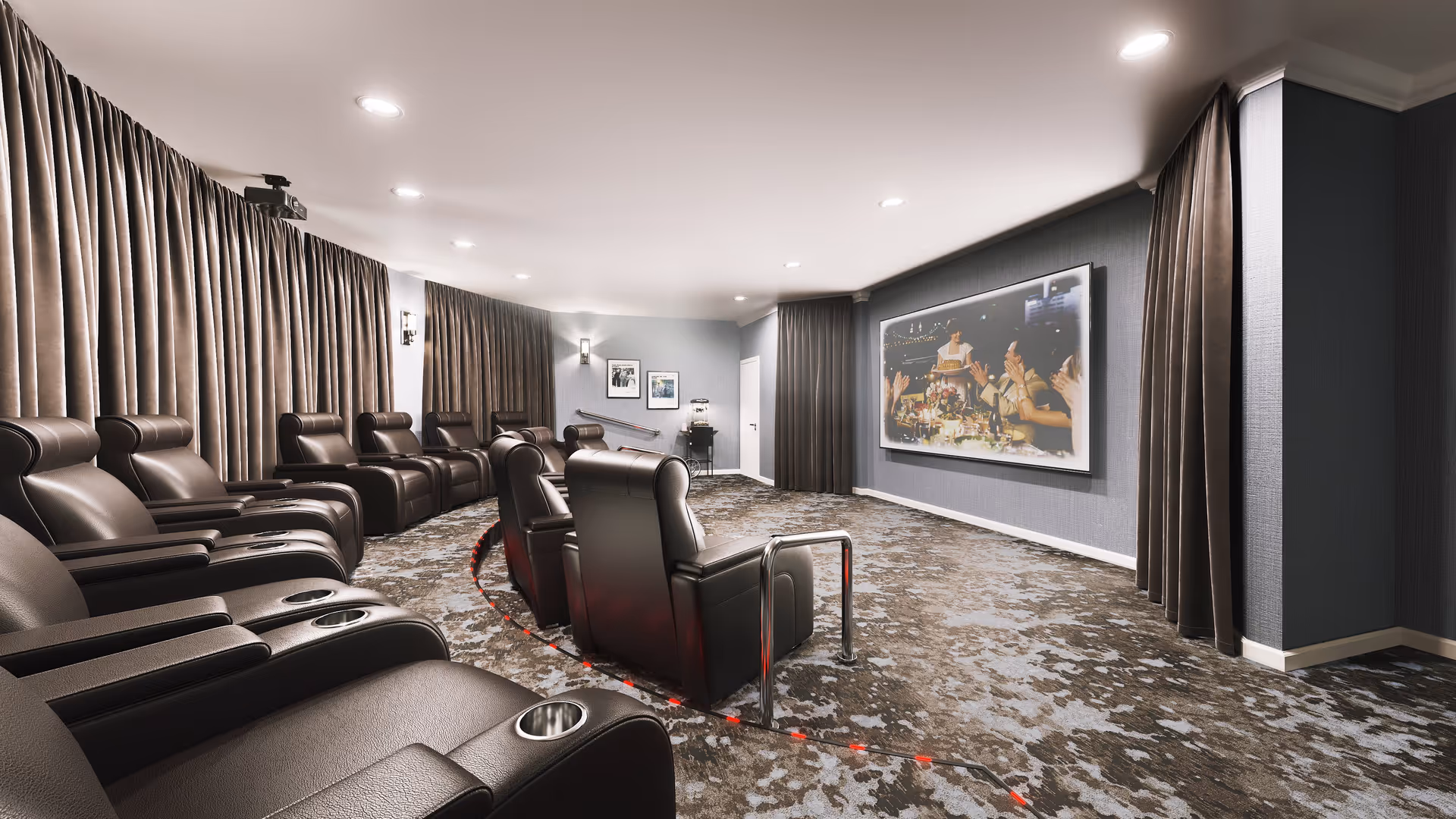 A modern home theater room with multiple rows of black leather recliner chairs facing a large screen on the wall. The room has dark curtains along the walls, recessed ceiling lights, and a patterned carpet floor. There is a popcorn machine and framed pictures on the back wall.